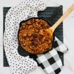 A cast iron skillet filled with baked beans and meat, perfect as one of the best burnt ends side dishes, sits on a black placemat. A wooden spoon rests in the skillet, with a polka dot towel and checkered oven mitt nearby.