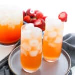 Two tall glasses of iced orange-pink drink, made with the Starbucks strawberry acai refresher recipe, each garnished with a strawberry, sit on a gray plate with a dark napkin. A matching pitcher and a bowl of strawberries are in the background.