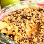 A glass pie dish filled with caramel apple cheesecake dip, topped with chopped nuts and chocolate chips; a bowl of sliced apples sits beside it, all resting on a colorful cloth.