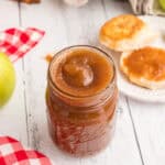 A mason jar filled with apple butter sits on a white wooden surface, surrounded by a green apple, cinnamon sticks, a red-checked cloth, and biscuits spread with apple butter—perfect for cozy fall breakfast ideas.