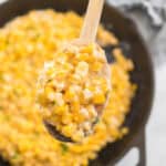 A wooden spoon holds a serving of creamy corn above a cast iron skillet filled with the same creamy corn mixture—an irresistible choice for sides for pulled pork sandwiches.