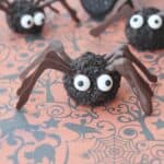 Three spider-shaped Halloween treats with candy eyes, chocolate legs, and a crumbly dark coating are arranged on an orange surface with black spooky patterns—perfect additions to your Halloween Dessert Table.