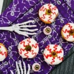 A cooling rack holds Halloween-themed cookies decorated as bloodshot eyeballs with white icing, red lines, and green candies. Plastic skeleton hands and fake eyeballs are on a purple skull-patterned cloth.