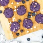 Purple blueberry cookies with white chocolate chips are arranged on a wooden board, surrounded by scattered blueberries and white chocolate chips. The background features a white and gray checkered cloth atop a marble surface.