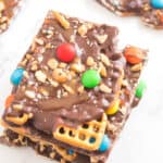 A stack of chocolate bark pieces topped with colorful candy-coated chocolates, pretzels, and chopped nuts sits on a white marble surface—perfect for festive New Years Eve desserts.