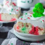 A slice of white cake with red and green spots, topped with whipped cream, festive sprinkles, and a green Christmas tree candy, sits on a stack of plates—an ideal treat inspired by classic 9x13 cake recipes for the holidays.