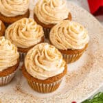 A plate of cupcakes topped with swirled cream frosting and sprinkled with cinnamon, surrounded by festive greenery and red berries on a holiday-themed table.