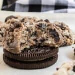 A close-up of Oreo cheesecake cookies dough with chocolate chunks, stacked on top of two chocolate sandwich cookies, with more cookie dough scoops in the background on parchment paper.
