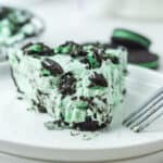 A slice of this no bake St Patrick's dessert, with creamy green and white filling, is topped with crushed mint Oreo cookies and served on a white plate with a fork. A partially visible pie and extra cookies appear in the background.