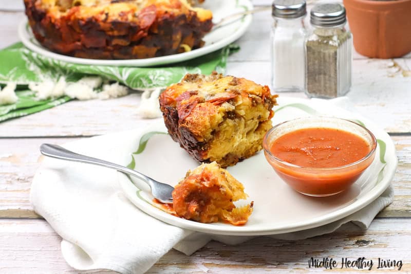 A slice of savory bread pudding, inspired by classic recipes with biscuits, sits on a white plate with a fork and a small bowl of marinara sauce, with more bread pudding in the background and salt and pepper shakers nearby.