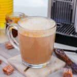 A glass mug of frothy caramel latte sits on a marble coaster with caramel candies, a swirl stick, and a coffee machine in the background—perfect inspiration for homemade Starbucks drink recipes. The setup is on a gray cloth.