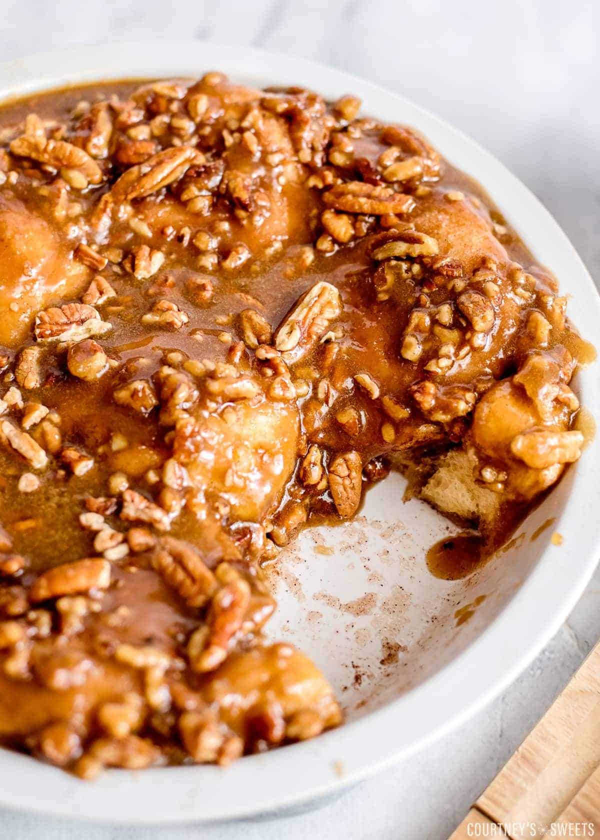 A round baking dish filled with sticky caramel pecan monkey bread made from biscuit dough, topped with chopped pecans and gooey sauce—a delicious Recipes with Biscuits treat, with a serving removed to reveal the soft bread inside.