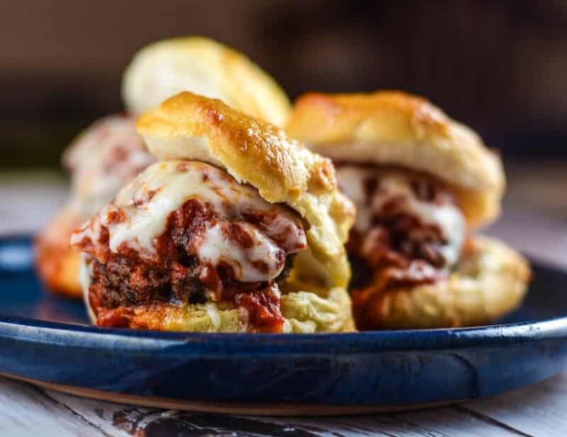 Three meatball sliders topped with melted cheese and marinara sauce are served on a blue plate. The sandwiches have golden, flaky buns and are arranged in a close-up, appetizing view.