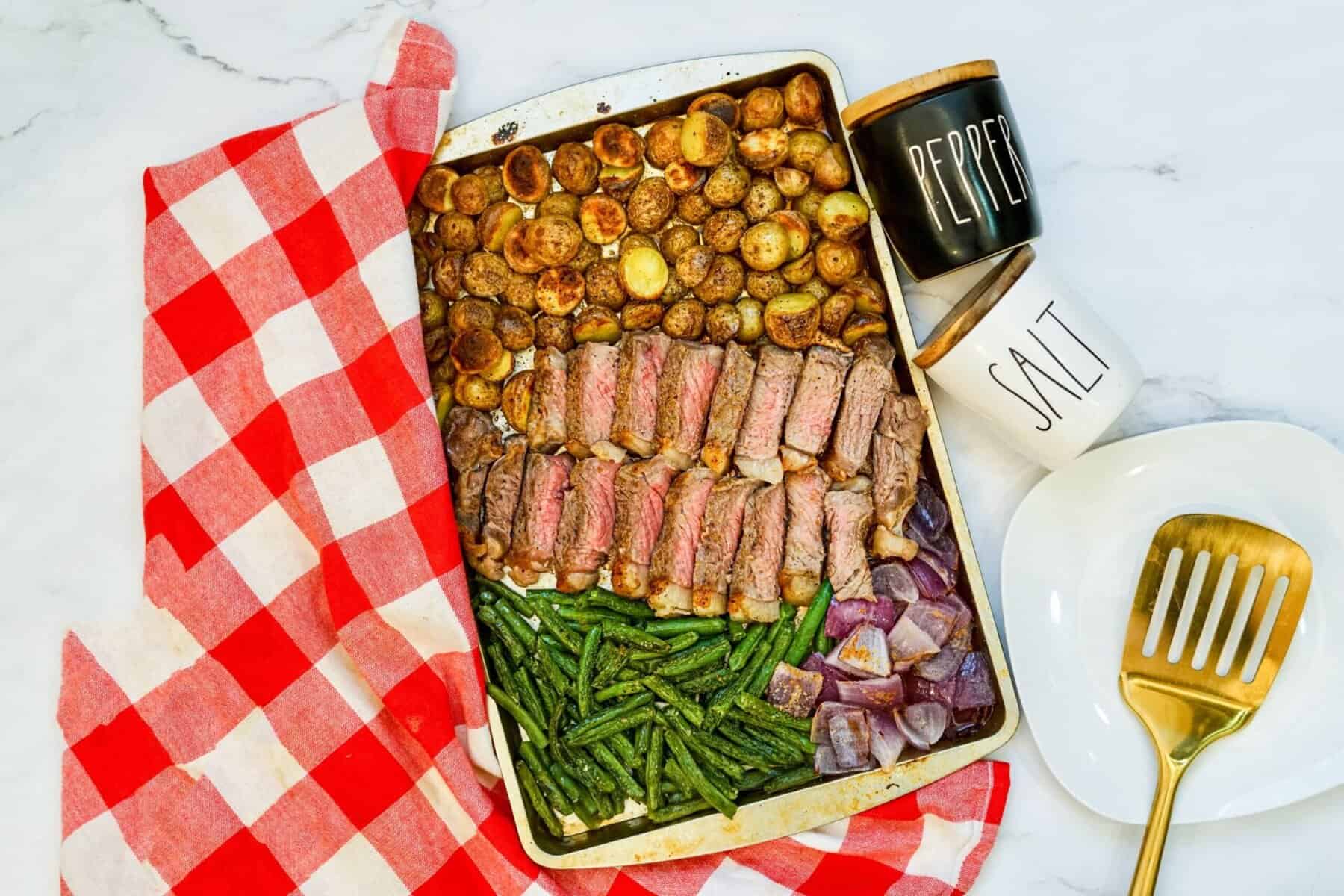 Overhead view of cooked steak slices, roasted potatoes, green beans, and red onion on a sheet pan. Salt and pepper containers on the side.