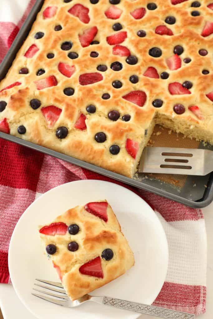 A baked sheet of pancake topped with blueberries and sliced strawberries, cut into squares.