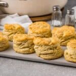 Eight golden-brown, flaky Pancake Mix Biscuits are arranged on a rectangular tray, with a white cloth, salt and pepper shakers, and stacked plates in the background on a gray surface.