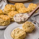 A wooden spoon holds creamy sausage gravy above a plate with two Pancake Mix Biscuits, with more biscuits and a pan of gravy in the background on a gray countertop.