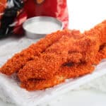 Crispy Dorito chicken tenders coated with a bright red, crunchy crust are arranged on a white plate with a small bowl of creamy dipping sauce and a bag of chips in the background.