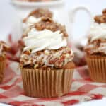 Three cupcakes topped with swirls of white frosting, chopped nuts, and a nut cluster are arranged on a red and white checkered cloth—an inviting addition to your easy Thanksgiving dessert recipes. A blurred white cup is visible in the background.