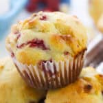A close-up of a freshly baked white chocolate raspberry muffin with visible fruit pieces in a white paper liner, stacked on another muffin. The golden-brown top hints at its soft, moist texture and delicious flavor.