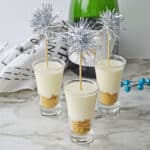 Three mini cheesecake champagne desserts in shot glasses, each with a crumb base and creamy topping, decorated with silver tinsel party picks. A champagne bottle, white cloth, and blue berries accent the marble surface.