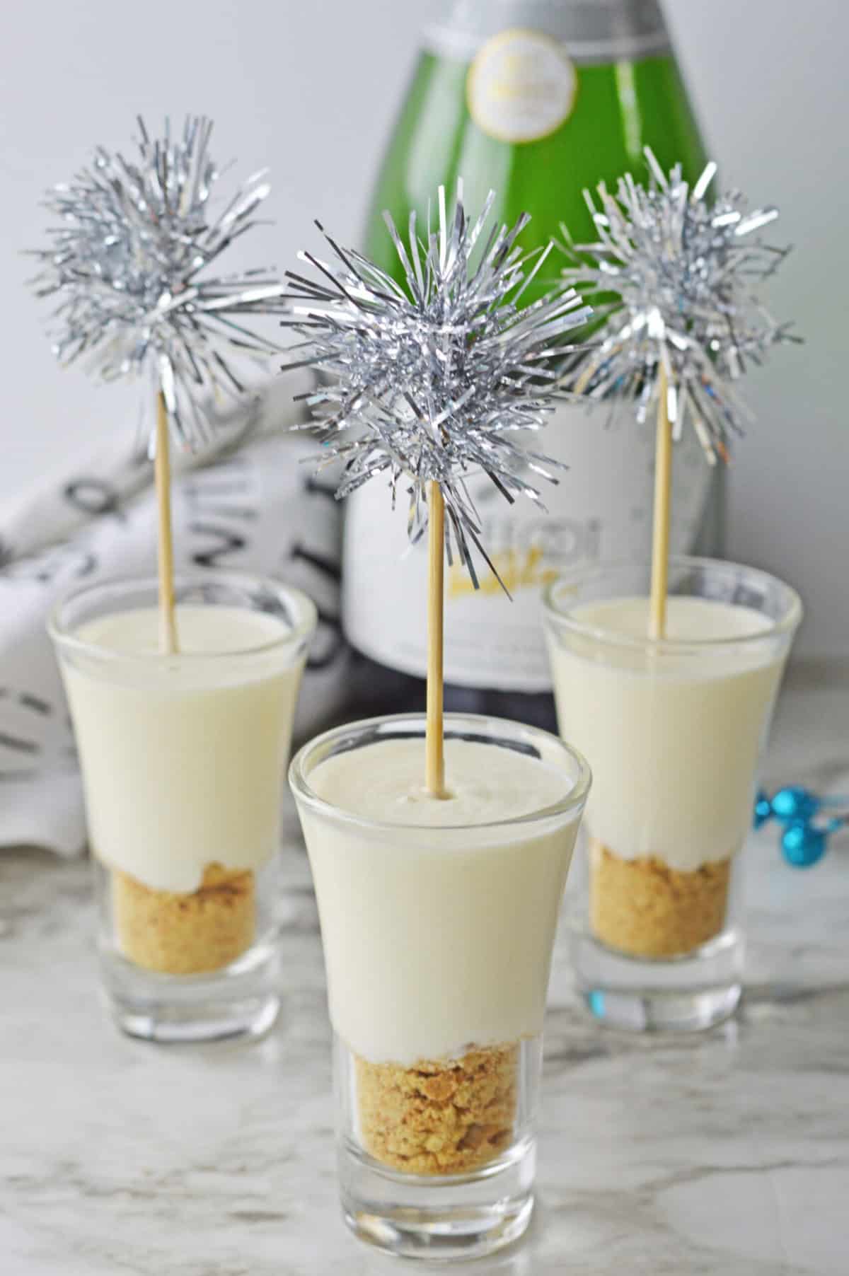 Three cheesecake shooters in shot glasses, each topped with a silver tinsel party pick. A bottle of sparkling wine and festive decorations add to the celebratory atmosphere in the background.