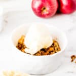 A white ramekin filled with air fryer apple crisp, topped with a scoop of vanilla ice cream, sits on a marble surface, with red and green apples in the background.