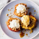 A plate with three air fryer baked apples filled with granola, topped with whipped cream and ice cream. One apple has caramel drizzle. A fork and napkin are beside the plate on a white surface.