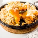 A close-up of a spoon lifting a serving of peach cobbler with a crumbly topping from a black baking dish, showing juicy peach filling and golden crust.