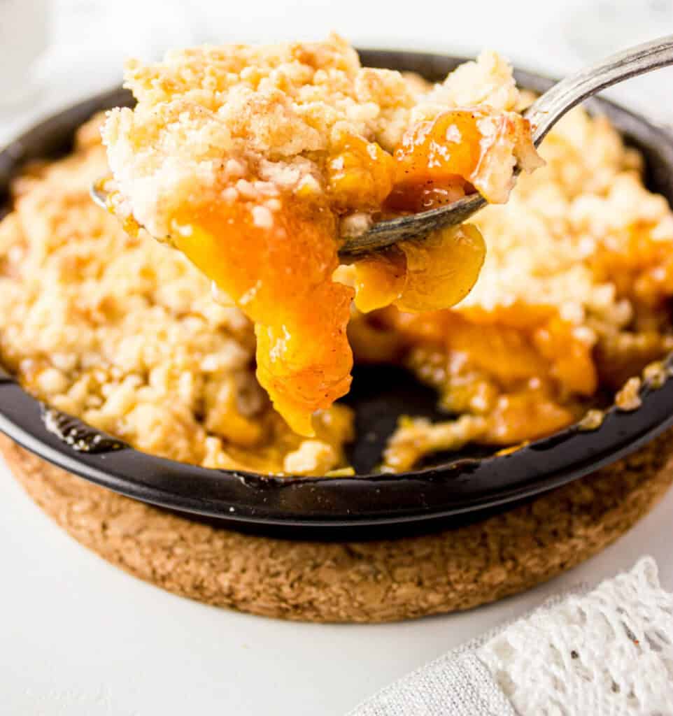 A close-up of a spoon lifting a serving of peach cobbler with a crumbly topping from a black baking dish, showing juicy peach filling and golden crust.