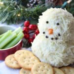 A snowman cheese ball is decorated with black peppercorns for eyes and mouth, and an orange nose. It’s surrounded by round crackers, celery sticks, and festive greenery in the background for a charming holiday touch.