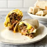A breakfast burrito sliced in half on a plate, showing sausage, eggs, bacon, cheese, and vegetables inside. A bowl of wrapped burritos sits in the background.