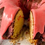 A bundt cake with a golden-yellow interior and a thick layer of glossy pink icing, shown with a large slice cut out to reveal the moist texture inside—perfect for those looking for festive Easter dessert ideas. Crumbs are scattered on the surface nearby.