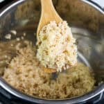 A wooden spoon holds a scoop of cooked instant pot brown rice over an open stainless steel electric pressure cooker containing more rice.