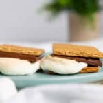 Two air fryer smores made with graham crackers, chocolate, and melted marshmallows sit on a blue plate, with an out-of-focus plant in the background and extra marshmallows nearby.