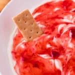 A close-up of a white bowl filled with creamy dip swirled with red strawberry sauce, with a rectangular graham cracker partially dipped in the mixture—perfect for showcasing this irresistible Strawberry Cheesecake Dip Recipe.