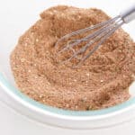 A metal whisk stirs a mixture of dry brown spices and visible seeds—perfect for Chili Seasoning—in a clear glass bowl on a white surface.