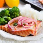 A dinner plate with a piece of grilled salmon fillet topped with thinly sliced red onions and a light glaze. The salmon is accompanied by steamed broccoli and a mound of fluffy white rice. In the background, fresh oranges and lemons are partially visible.