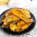 Plate of baked potato wedges on a printed cloth with garlic and cheese in the background.