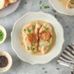 A plate of lemon chicken Francaise featuring two lightly breaded chicken breasts topped with slices of caramelized lemon and a light, glossy sauce. Fresh chopped parsley is sprinkled on top, and a silver fork rests across the chicken. The dish is served on a white scalloped-edge plate.