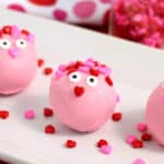 Three pink, round Valentine’s Day Oreo Balls decorated with candy eyes and colorful heart-shaped sprinkles sit on a white plate, creating a cute, playful treat.