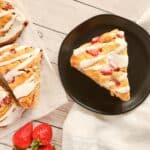 A triangular strawberry scone with white icing sits on a black plate next to more scones on parchment paper and fresh strawberries, making it perfect for valentine breakfast ideas on a white wooden surface with a white cloth nearby.