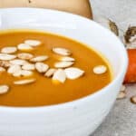 A close-up of a white bowl filled with creamy orange soup, garnished with pumpkin seeds. In the background, a carrot, garlic bulb, and part of a pumpkin are visible on a light surface—perfect inspiration for delicious soup recipes.