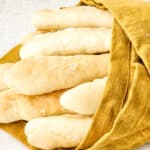 A group of golden-brown breadsticks, made with an Olive Garden breadsticks recipe and sprinkled with coarse salt, partially wrapped in a mustard yellow cloth on a light textured surface.