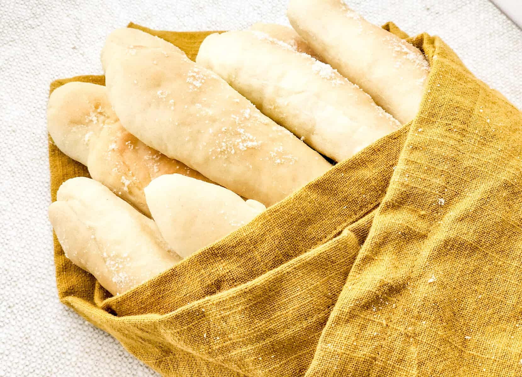 A bundle of golden breadsticks, just like the Olive Garden breadsticks recipe, sprinkled with salt and wrapped in a mustard yellow cloth, resting on a light textured surface.
