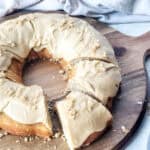 A vanilla bundt cake with light brown frosting and sprinkled crumbs sits on a wooden board. Several slices are cut, revealing the cake’s soft interior. A white towel and knife are partially visible in the background.