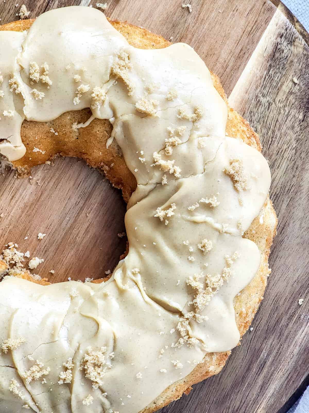 A close-up of a vanilla bundt cake with creamy beige frosting and sprinkles of brown sugar on top, placed on a wooden board.