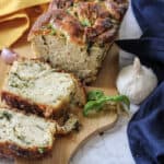 A sliced, homemade herb bread loaf sits on a wooden board with fresh basil and garlic bulbs nearby—an inviting choice for easy appetizers for a party—surrounded by yellow and blue cloths on a marble surface.
