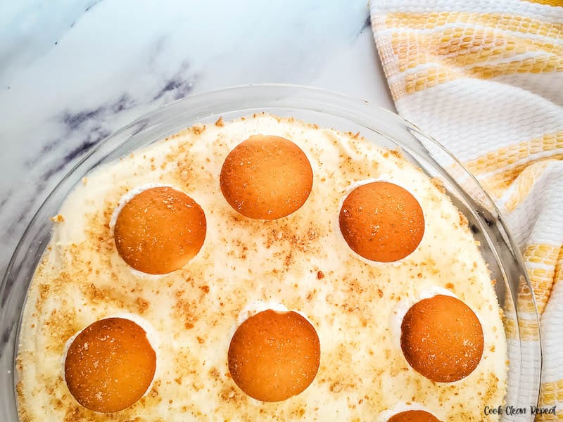 A close-up of a banana pudding dessert topped with round vanilla wafer cookies and layers of creamy Cool Whip Desserts in a glass dish, with a yellow and white checkered towel beside it on a marble countertop.