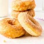 A stack of four homemade donuts coated in cinnamon sugar sits on a white surface, with a bottle of milk and a purple-striped straw blurred in the background.